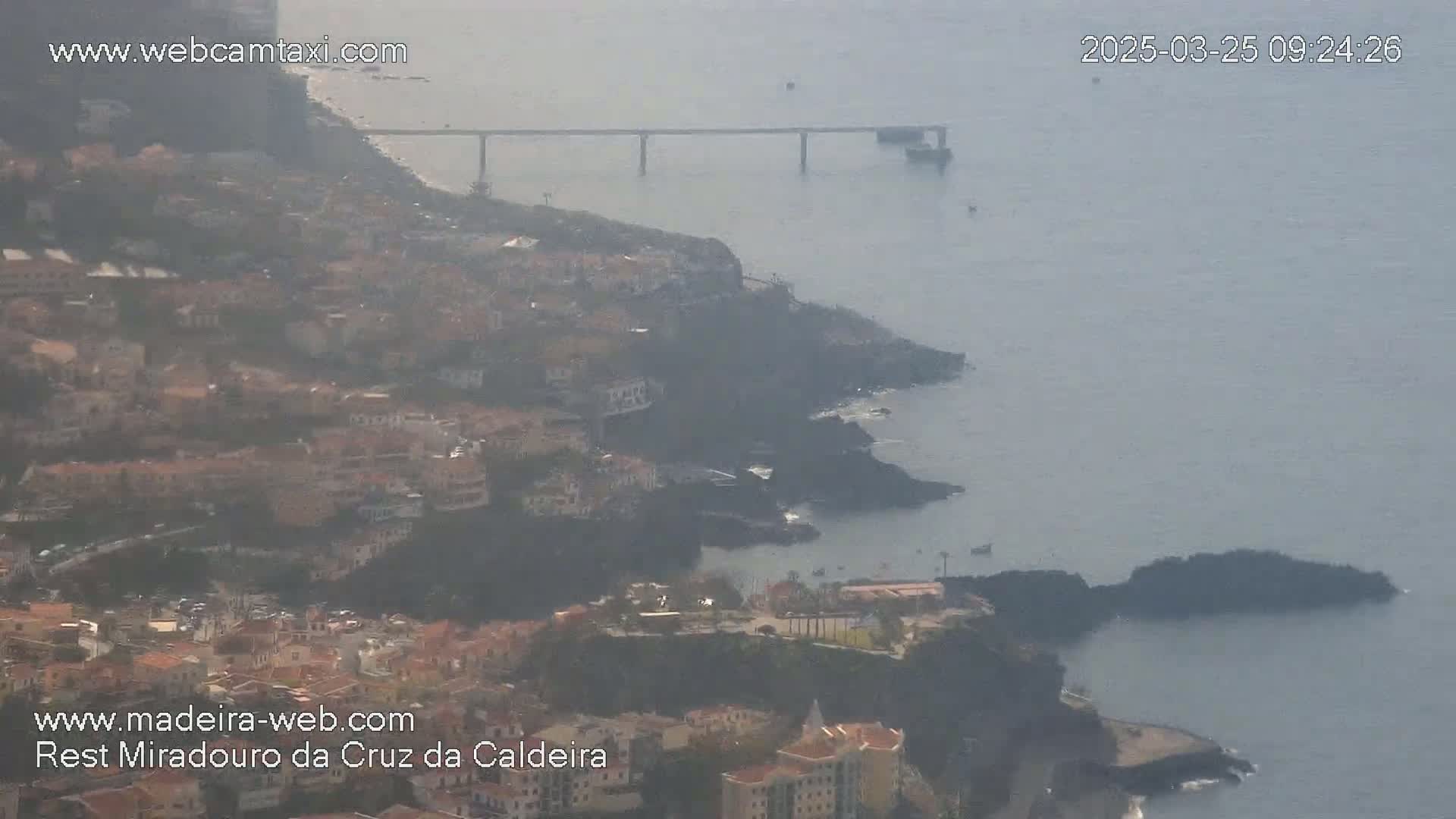 A hazy coastal town clings to a rocky hillside overlooking a calm ocean, with a long bridge visible in the distance.