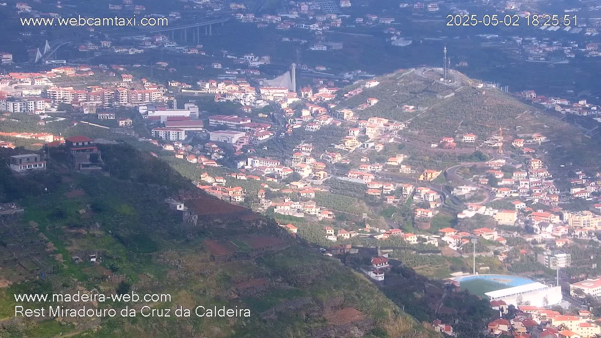 A hillside town with red-roofed houses, terraced farmland, and a distant bridge is partially shaded on a sunny day.