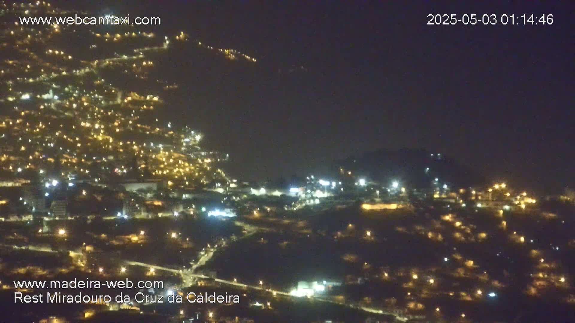 A nighttime, aerial view shows a hillside town illuminated by numerous lights under a dark, hazy sky.