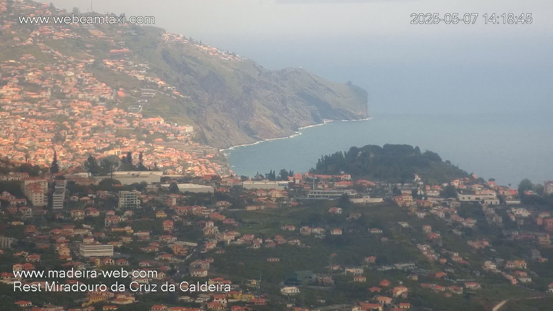 A hazy coastal town sprawls across a hillside, descending toward a calm ocean meeting a steep cliff.