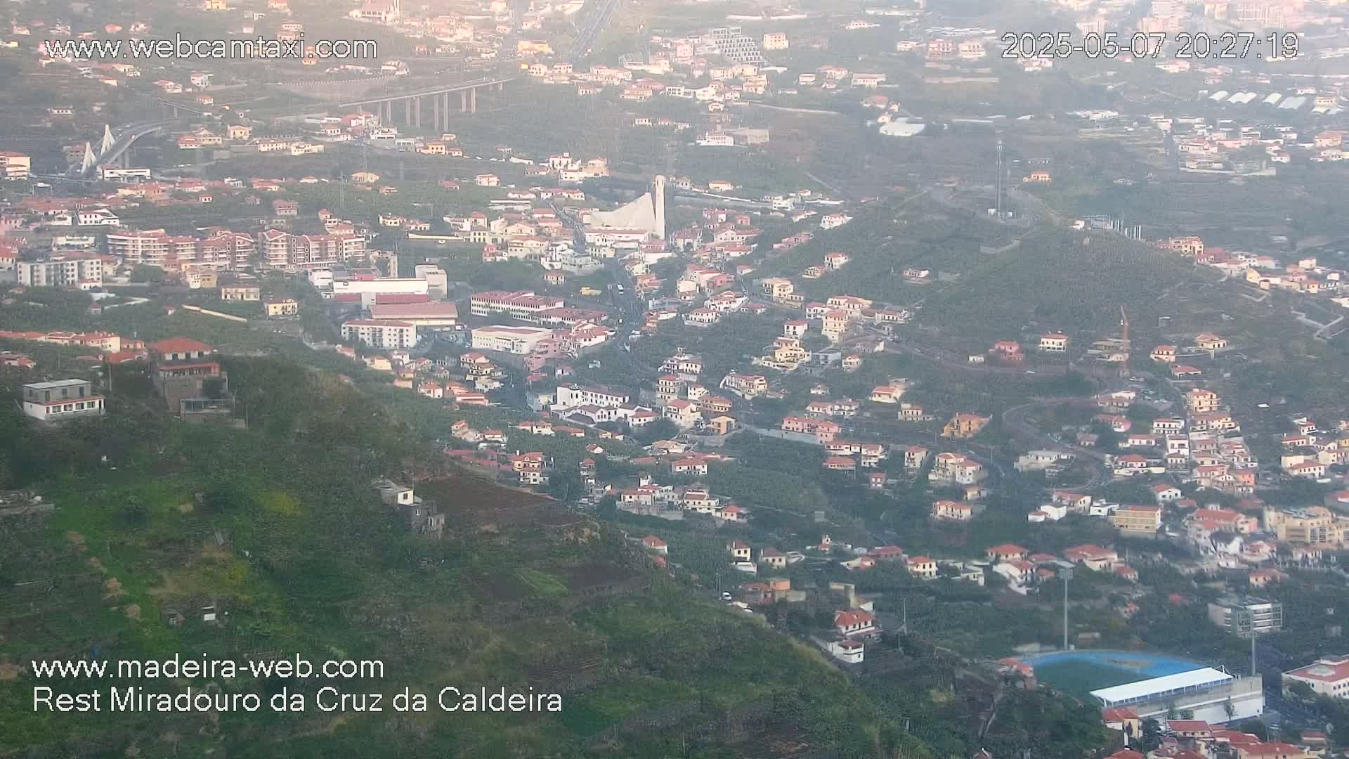 A hazy, high-angle view shows a hillside town with many small buildings, a large stadium, and a bridge in the distance.