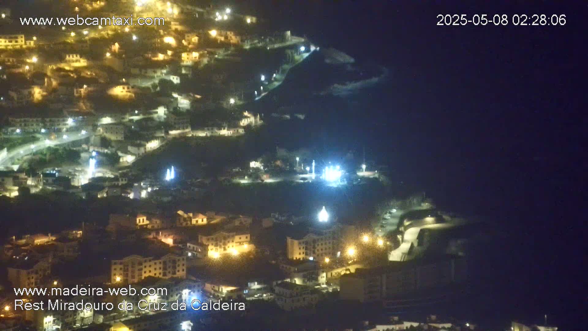 A nighttime, high-angle view of a hillside town illuminated by streetlights, with the dark ocean visible in the background.