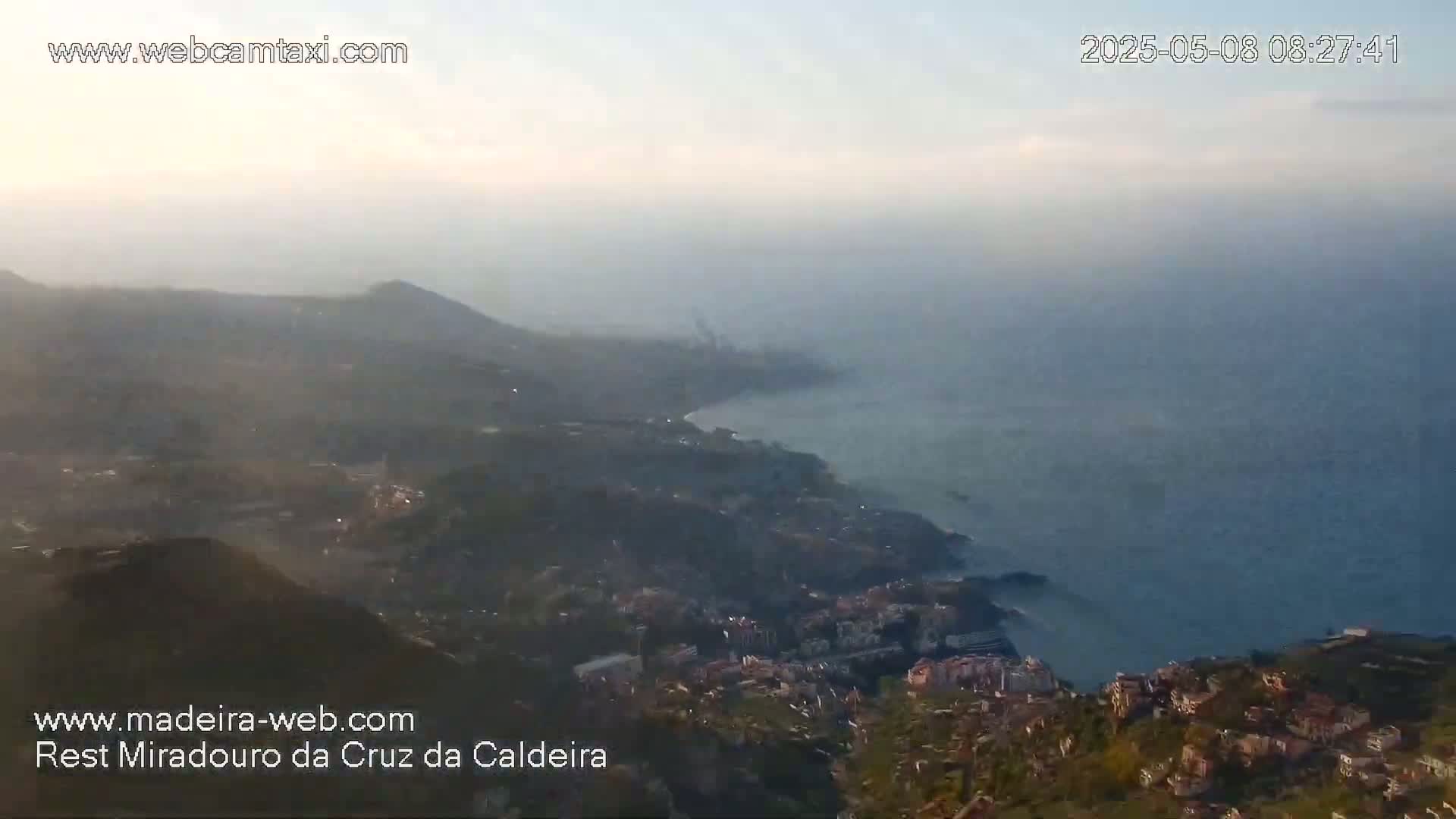 A hazy, sunrise view shows a coastal town nestled into hills overlooking a calm ocean.