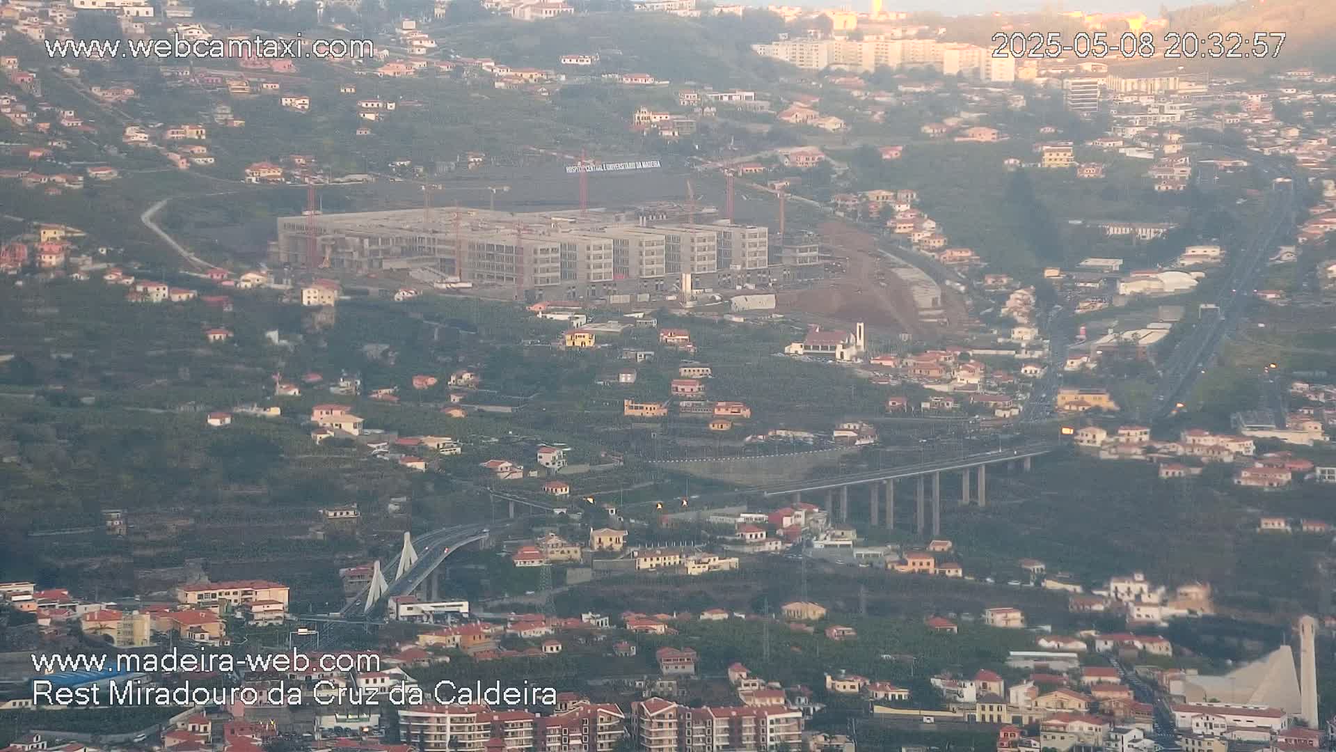 An aerial view shows a large building under construction, a long elevated highway, and numerous houses nestled on a hillside in hazy, possibly evening light.