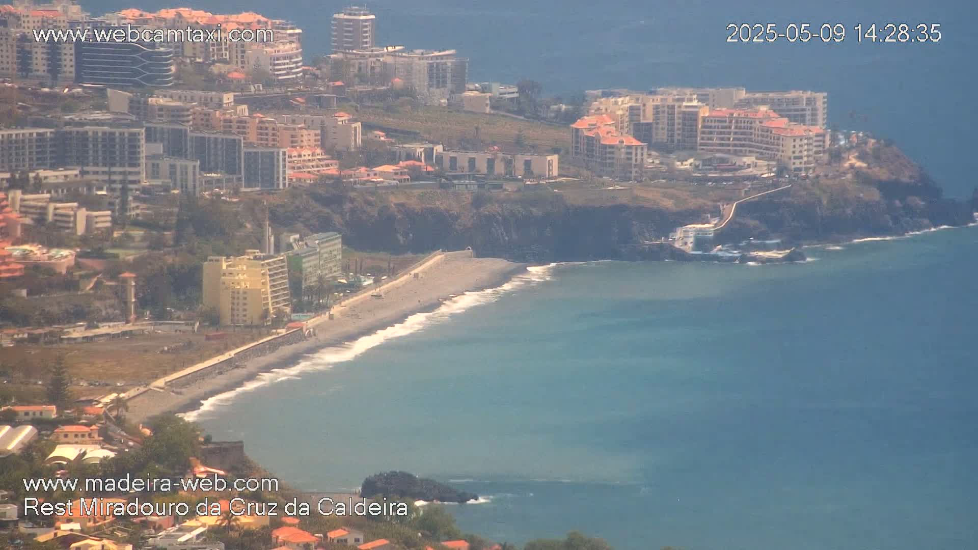 A coastal town with numerous buildings sits on a cliff overlooking a beach with gentle waves under a hazy sky.