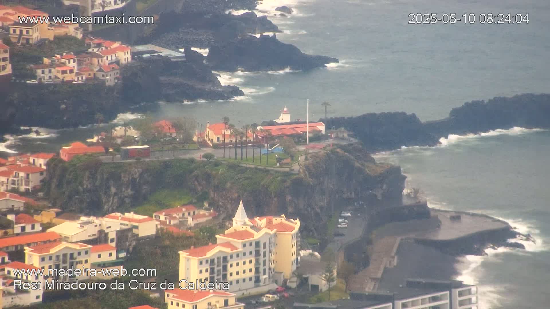 A coastal town with red-roofed buildings is nestled on a cliff overlooking a dark-grey ocean with moderate waves, under an overcast sky.