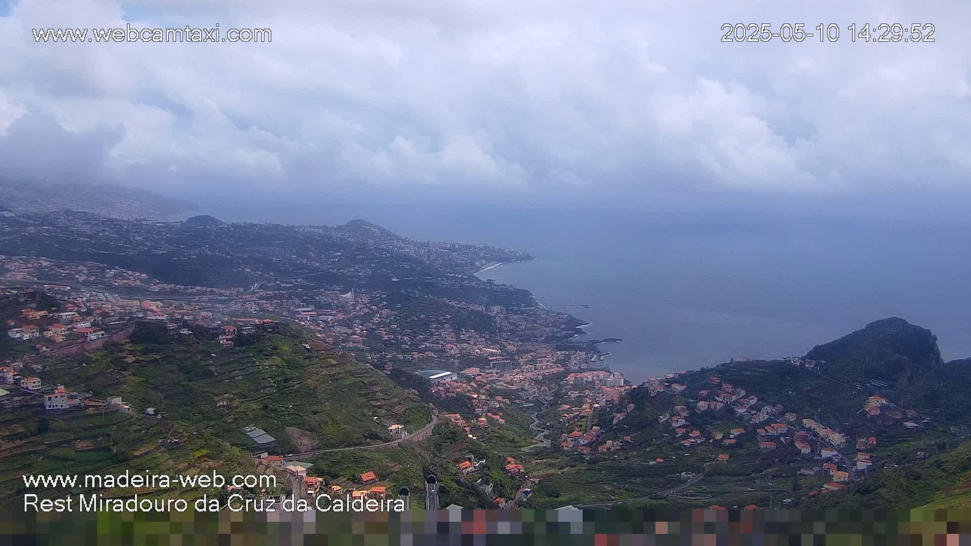 A coastal town nestled in a mountainous region is visible under a mostly cloudy sky.