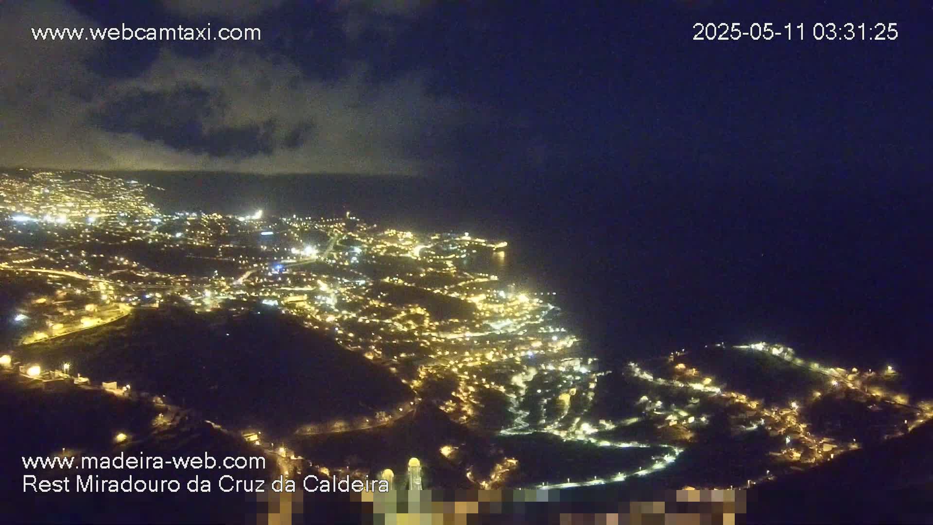 A nighttime, aerial view of a city illuminated by streetlights, nestled along a dark coastline under a partly cloudy sky.