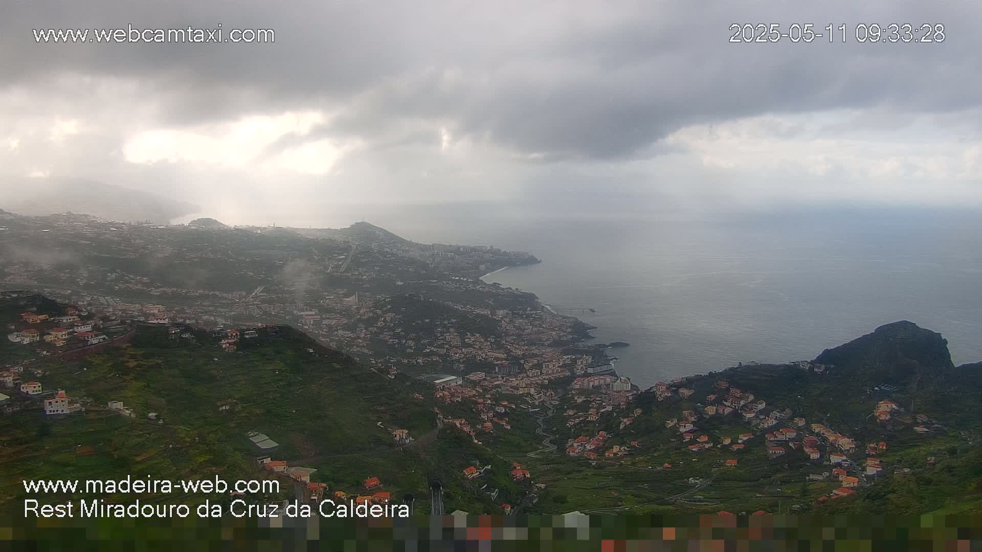 A coastal town nestled in a mountainous region is viewed from above on an overcast day, with the ocean visible in the distance.
