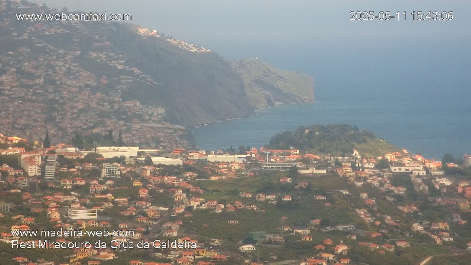 A hazy coastal town with red-roofed houses sprawls across a hillside, overlooking a calm ocean and a steep, green cliff.
