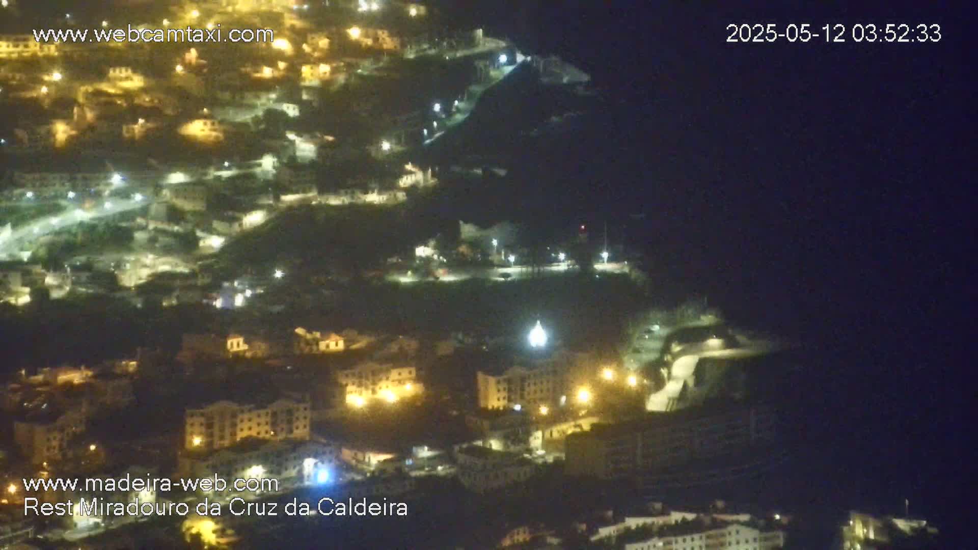 A nighttime, aerial view of a hillside town illuminated by streetlights, with a dark ocean visible in the background.
