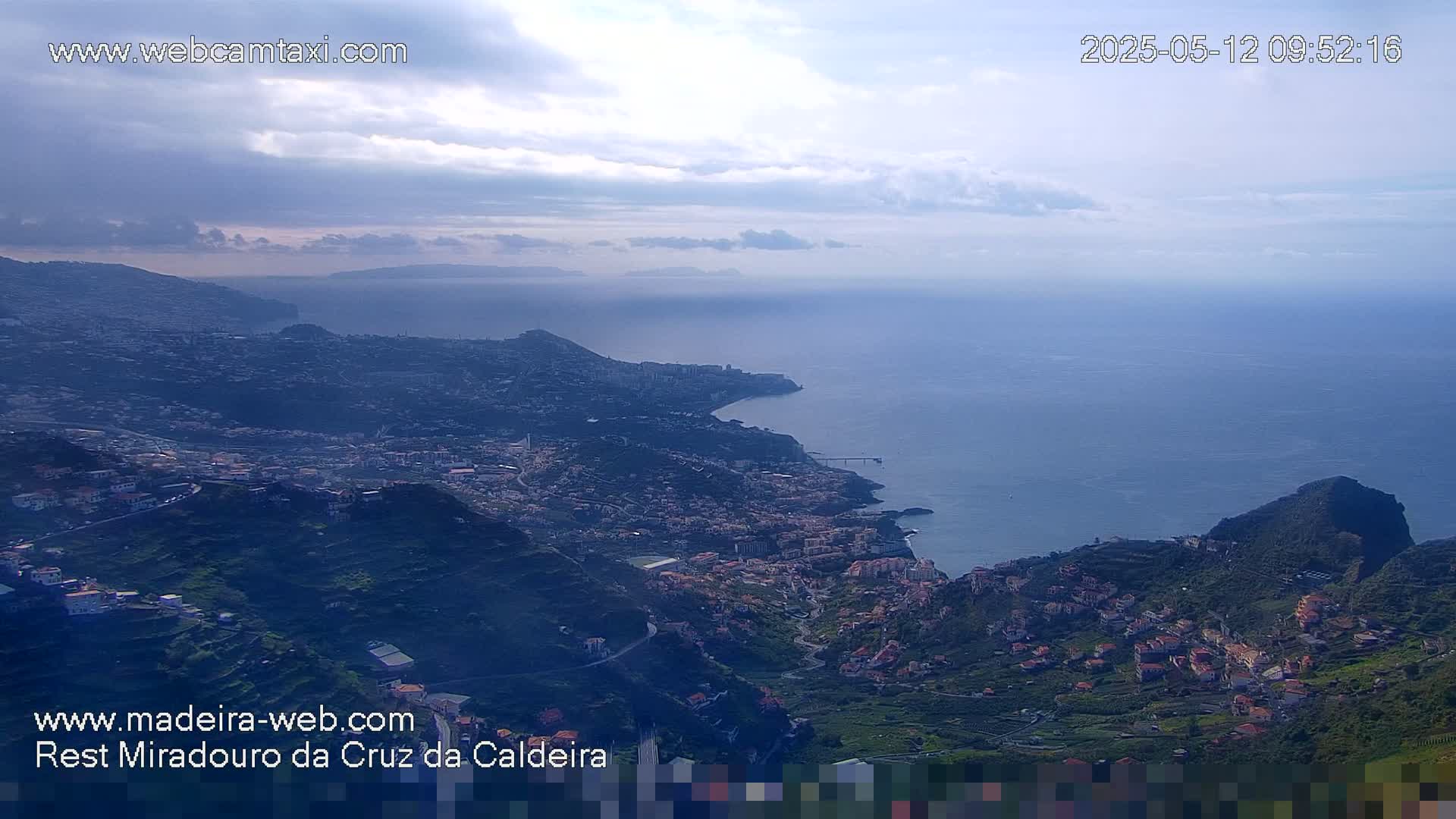 A coastal town nestled in a mountainous region overlooks a calm ocean under a partly cloudy sky.