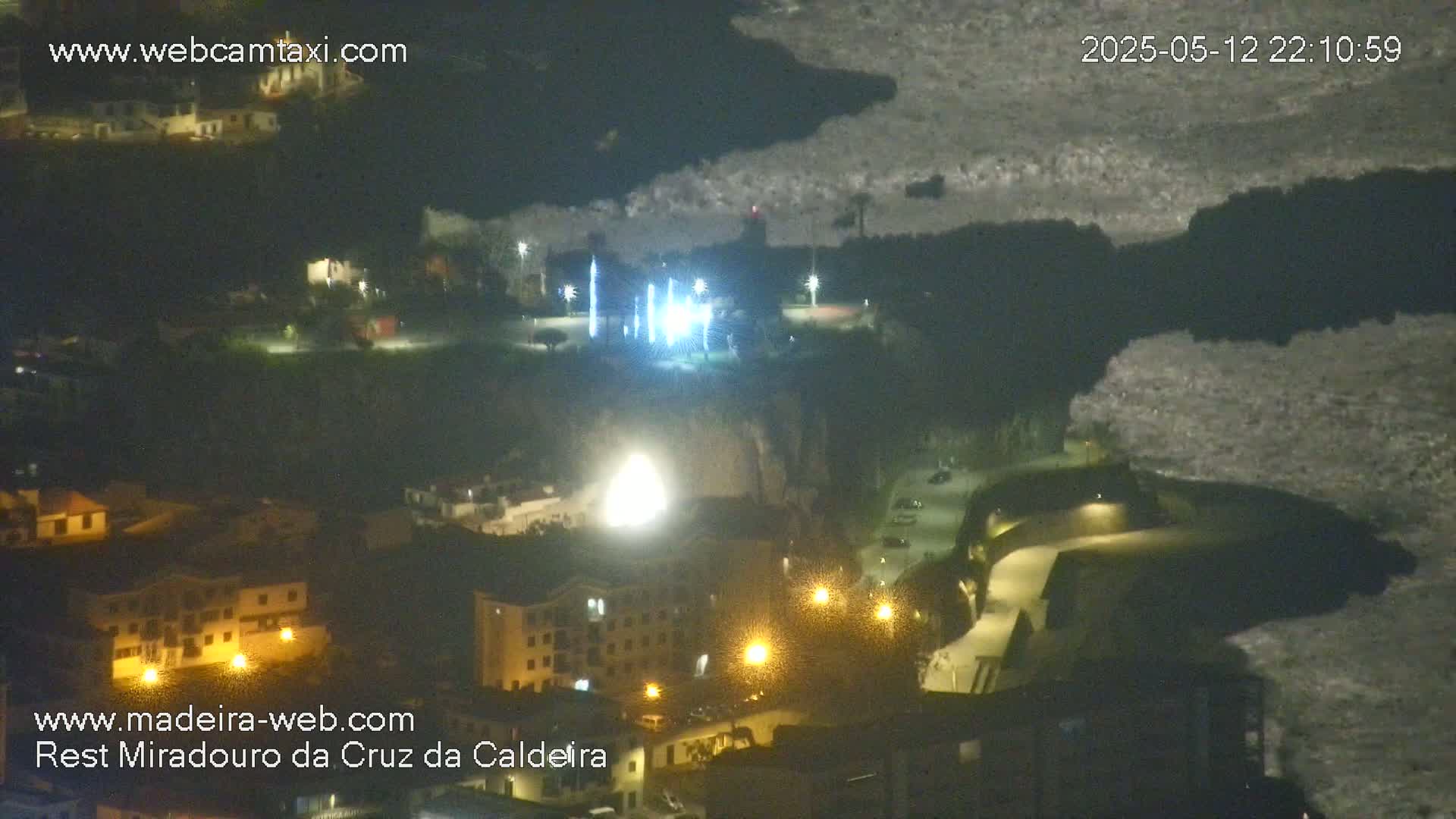 An aerial nighttime view of a town situated on a cliff overlooking a dark body of water under a cloudy sky.