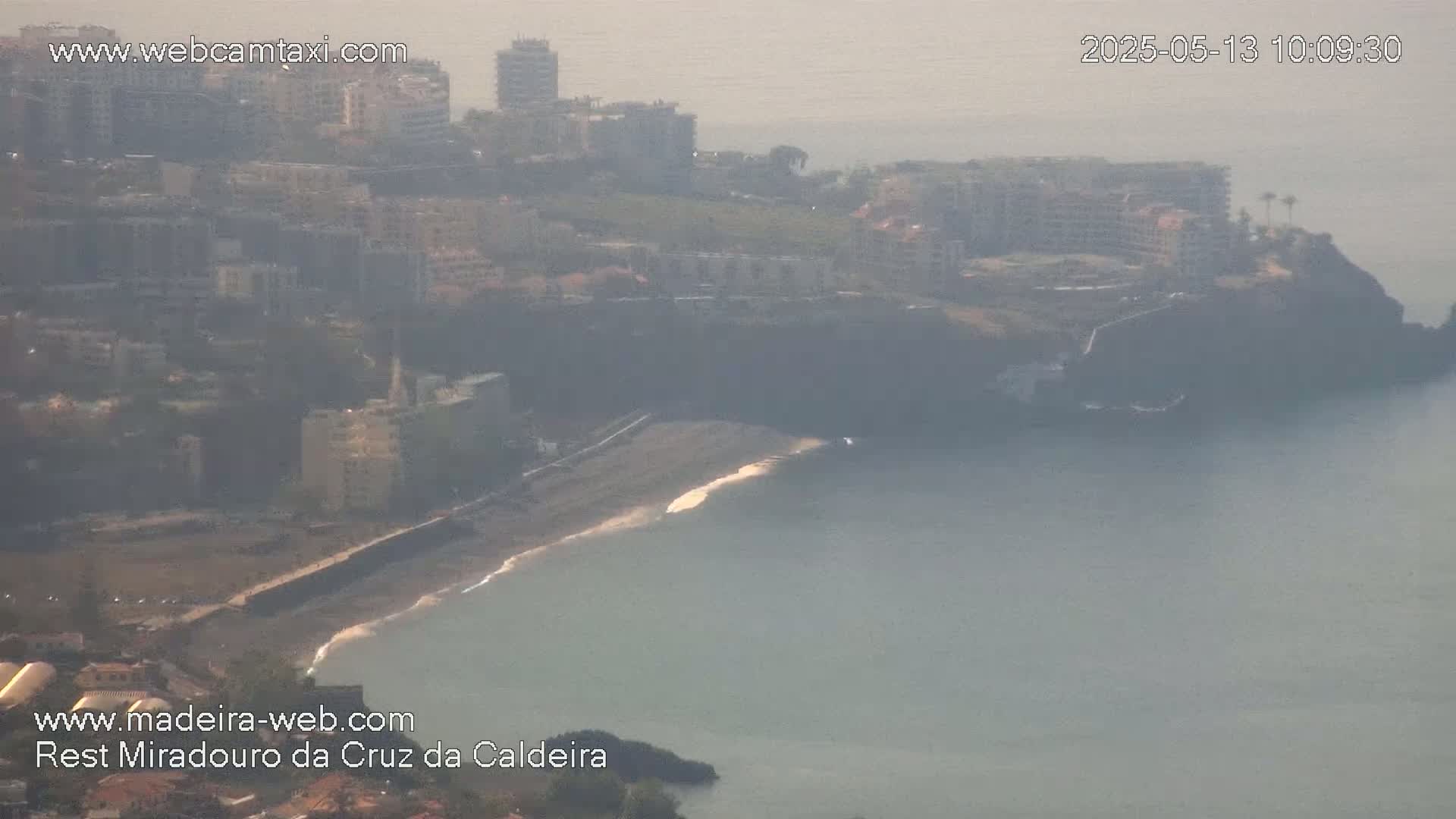 A hazy coastal scene shows a dark-colored beach curving into calm water near buildings on a cliff.
