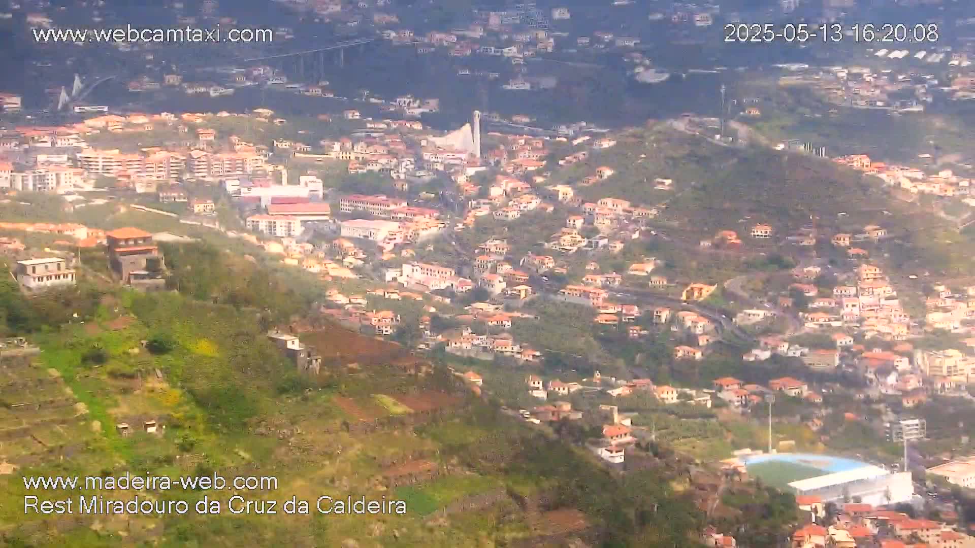 A hillside town with many low buildings and a soccer field is visible from above on a hazy day.