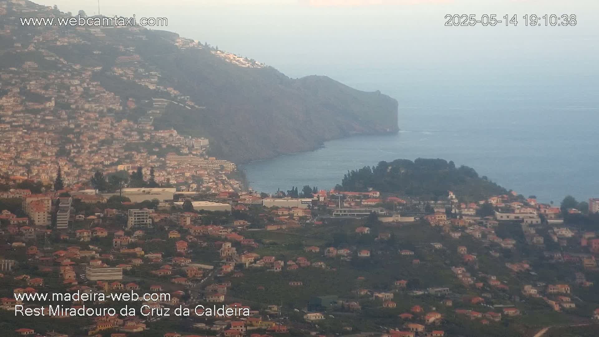A hazy coastal town nestled on a hillside overlooking a calm ocean, with a steep cliff in the background.