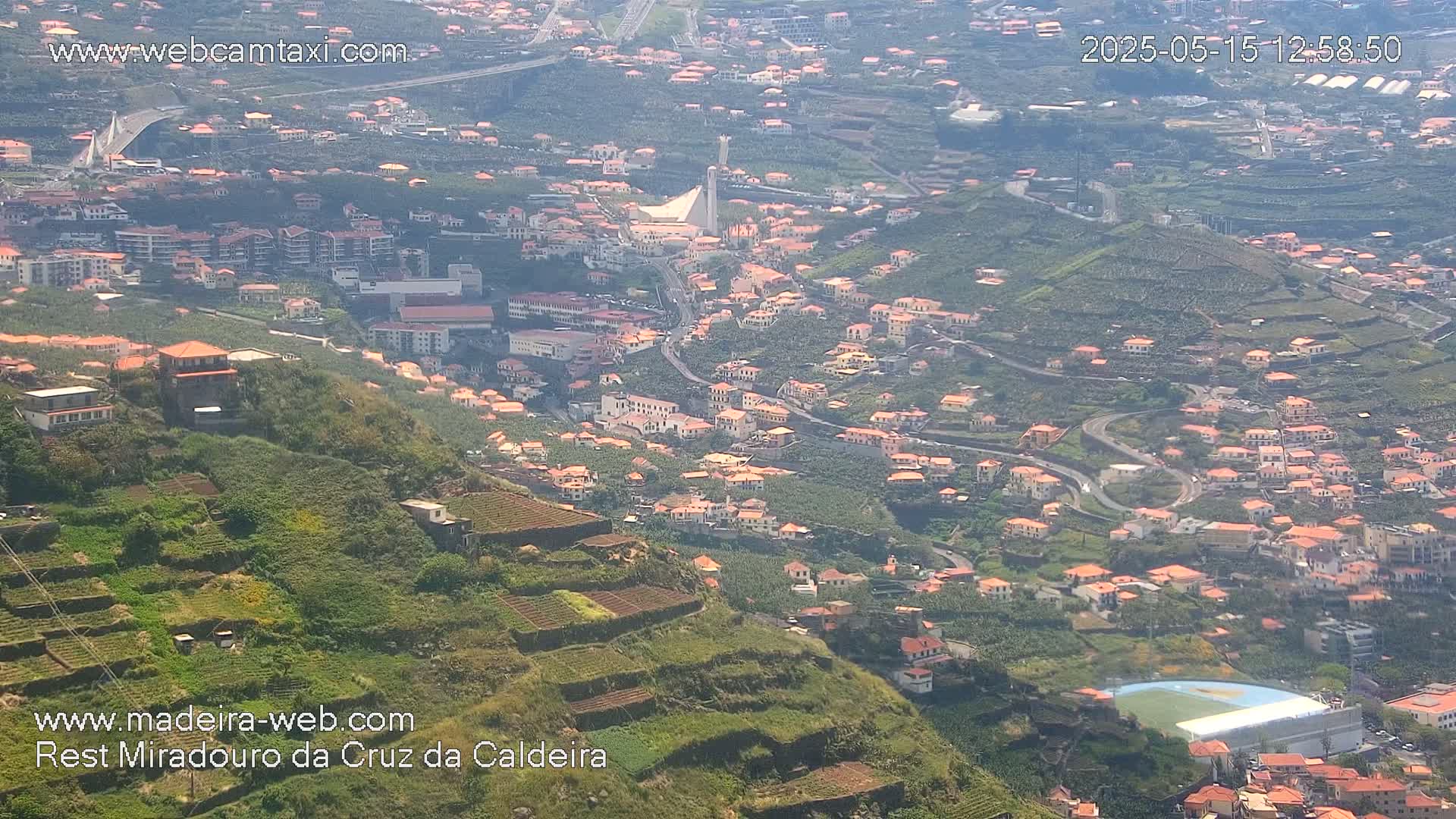 A hillside town with red-roofed buildings and terraced farmland is visible on a sunny day, overlooking a valley and a bridge in the distance.