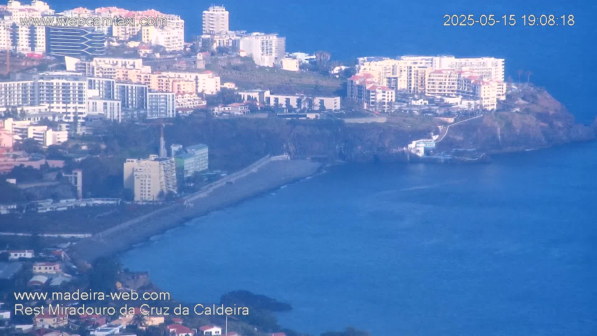 A coastal city with numerous buildings is situated on a cliff overlooking a calm, blue ocean.