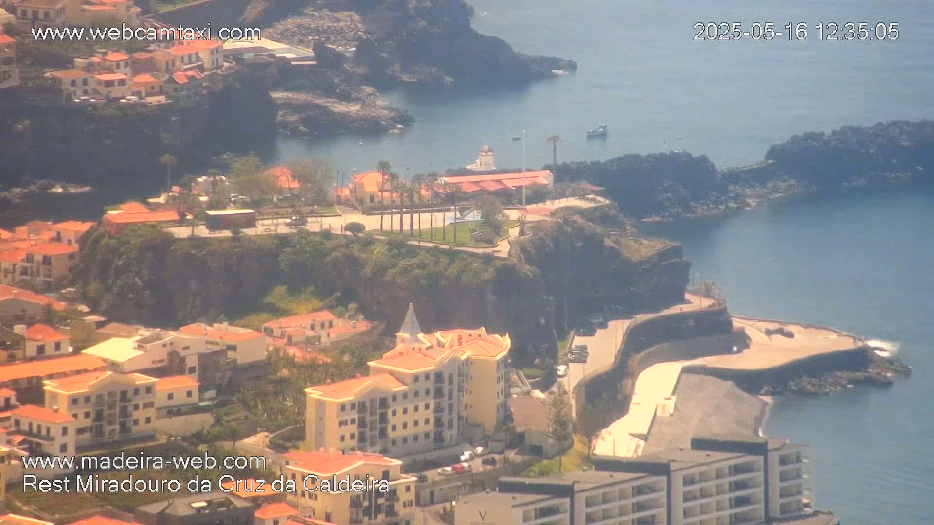 A coastal town with red-roofed buildings clings to a cliffside overlooking a calm ocean on a sunny day.