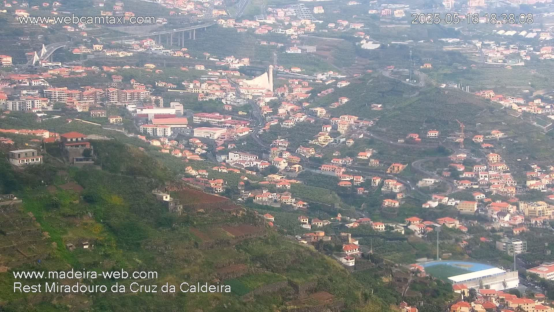 A hazy, high-angle view shows a sprawling hillside town with numerous terracotta-roofed buildings, terraced farmland, and a large sports field, all overlooking a long bridge and distant ocean.