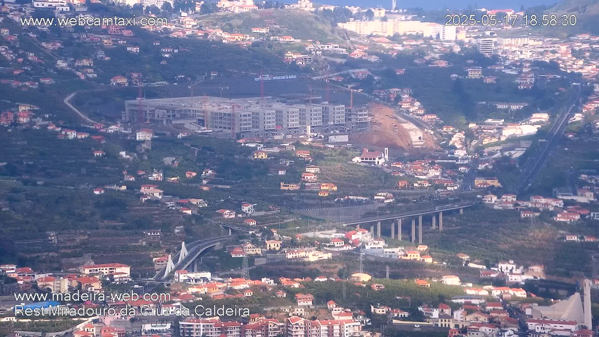 An aerial view shows a hillside town with a large building under construction and a long elevated highway.