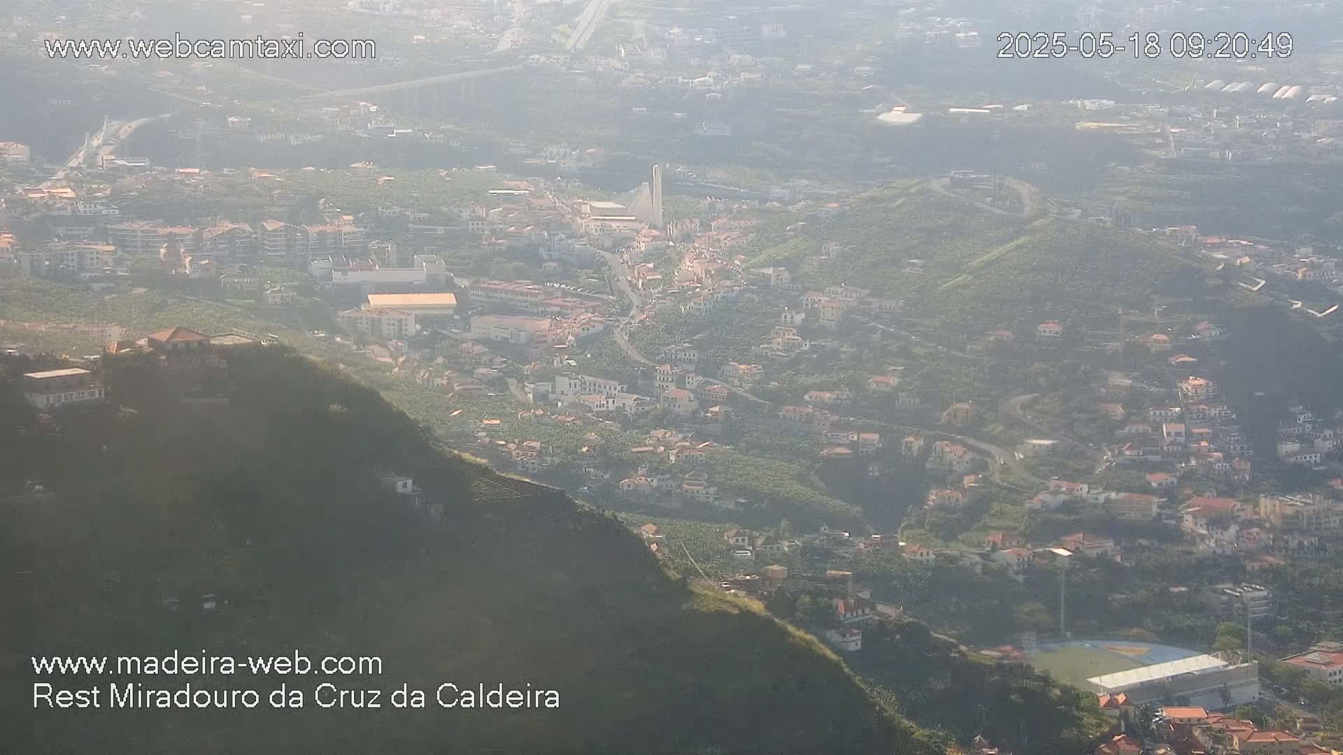 A hazy, high-angle view shows a town nestled in a hillside, with buildings scattered among green vegetation and a playing field visible in the lower right.