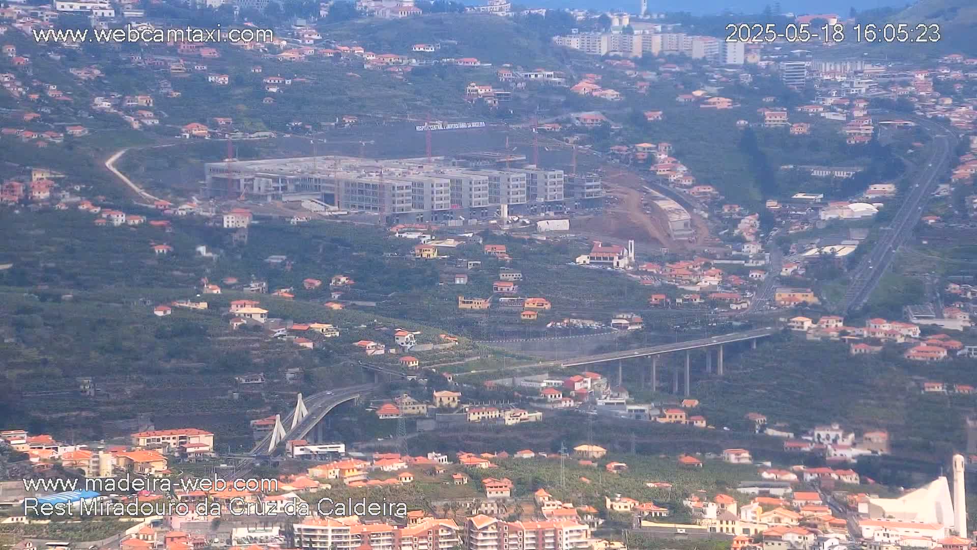 A high-angle, long shot shows a coastal town with a large building under construction, multiple bridges, and a highway under an overcast sky.