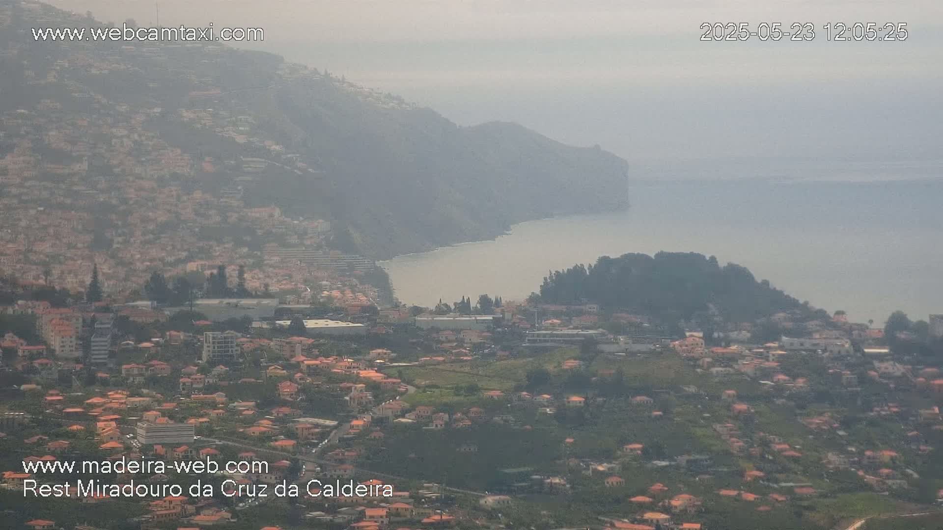 A hazy coastal town nestled on a hillside overlooking a calm ocean, with a large cliff rising from the water in the background.