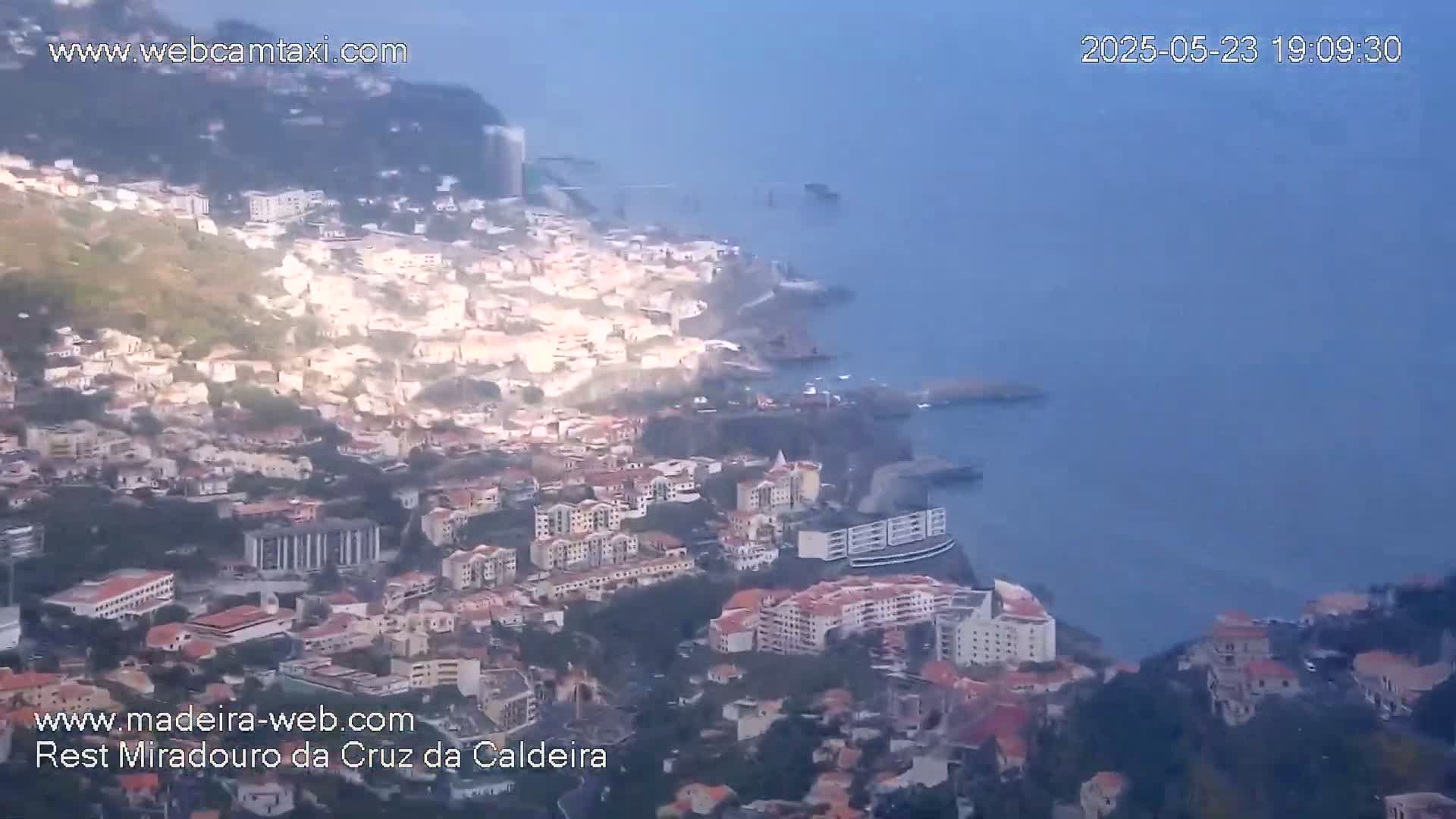 A hazy, high-angle view shows a coastal town built into a hillside, descending to a calm ocean.