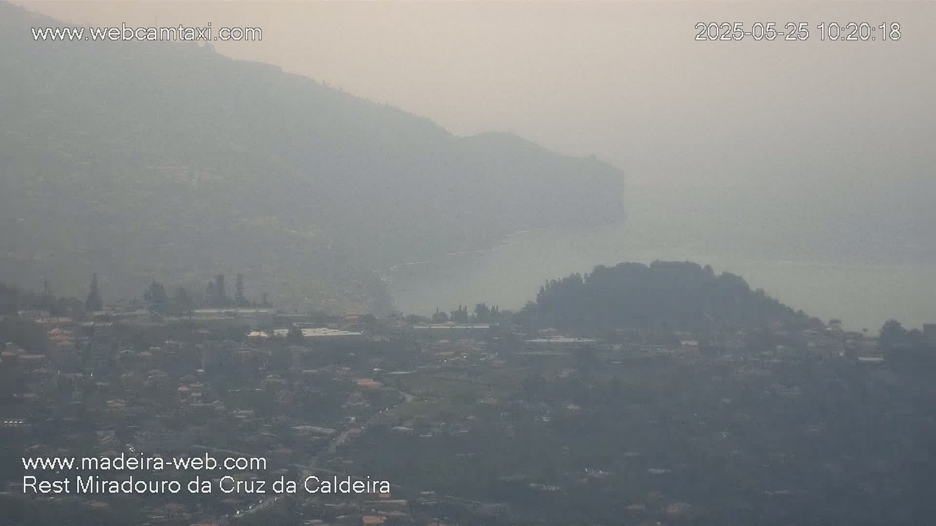 A hazy view shows a town nestled between hills and a body of water under a hazy sky.