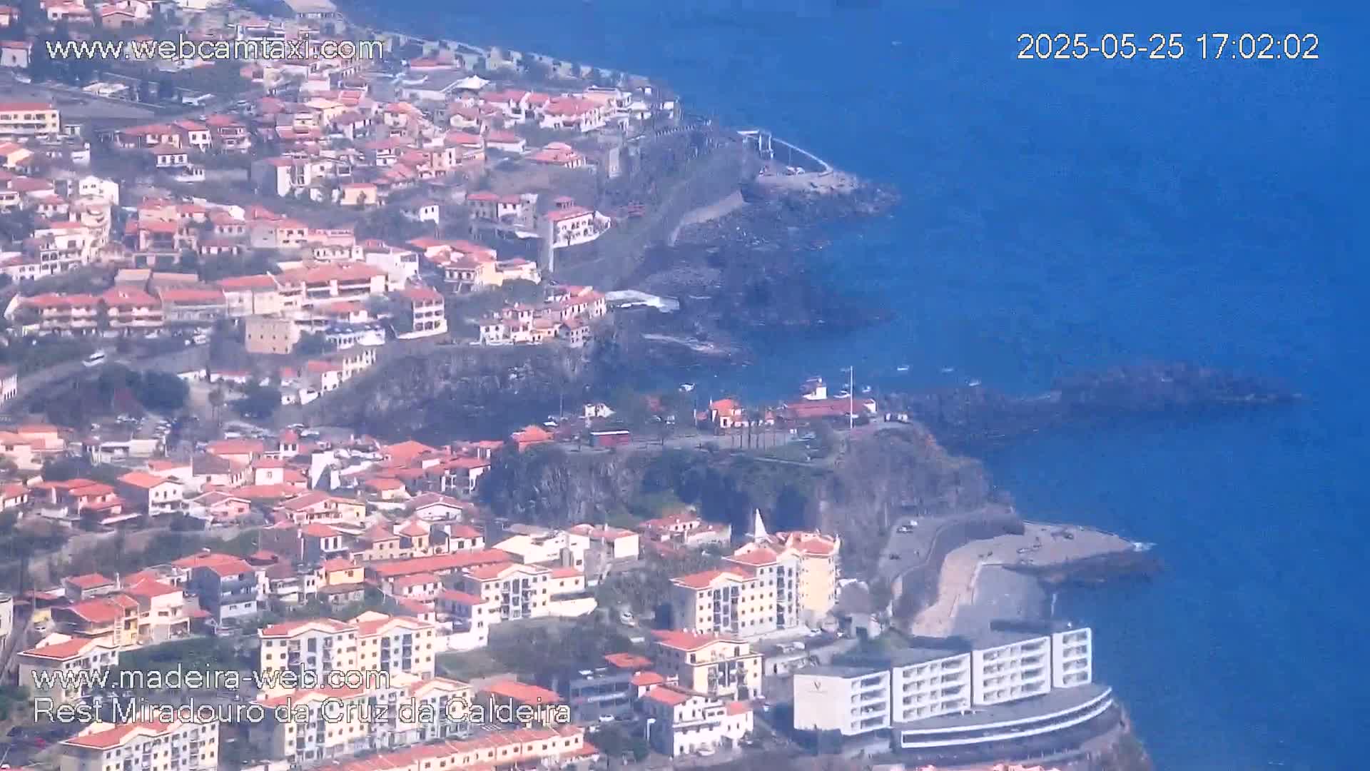 A coastal town with red-roofed buildings clings to a cliffside overlooking a calm, blue ocean.