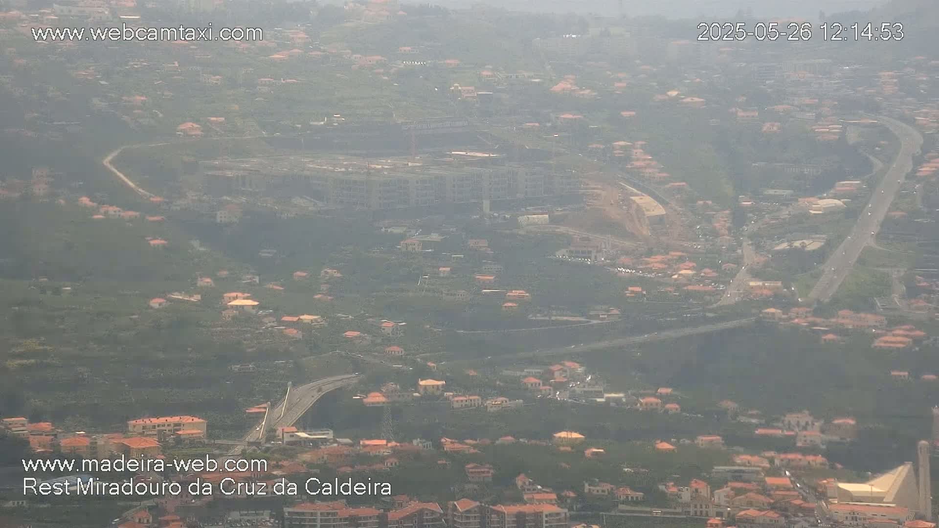 A hazy view shows a hillside town with numerous light-brown buildings and winding roads, a large construction site, and a highway.