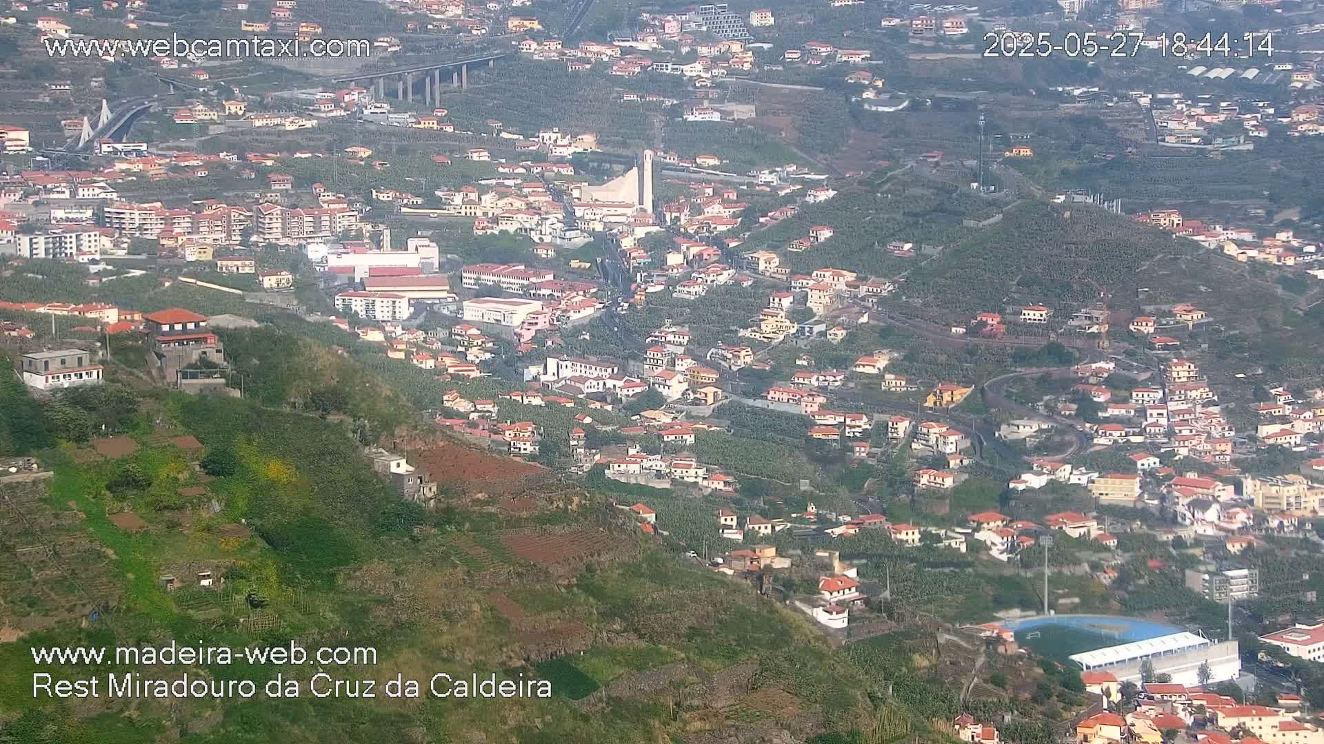 A high-angle, long shot shows a hillside town with red-tiled roofs, terraced farmland, and a sports field, under hazy daylight conditions.