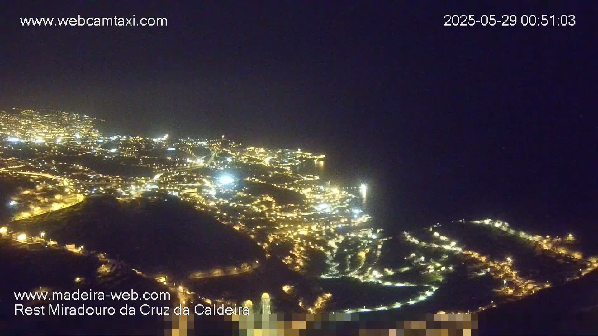 A nighttime, aerial view shows a city illuminated by many lights, extending down a hillside toward a dark ocean.