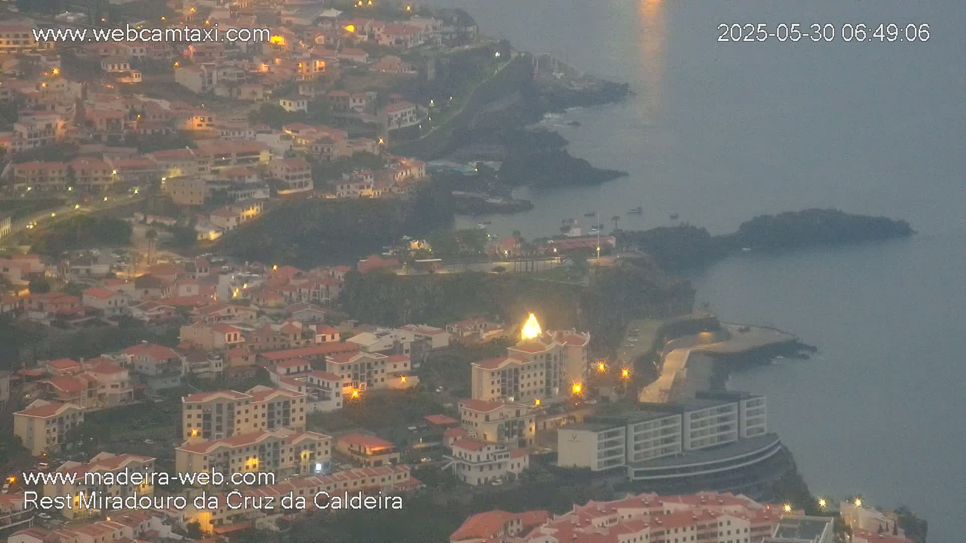 A coastal town at twilight is partially obscured by haze, with its lights illuminating buildings and streets that slope down toward the dark ocean.