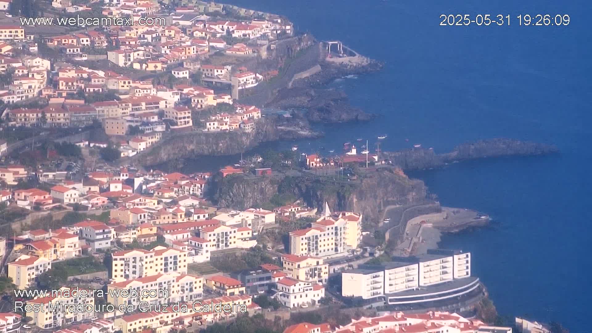 An aerial view shows a coastal town built on a hillside, with buildings cascading down towards a dark blue ocean under a sunny sky.