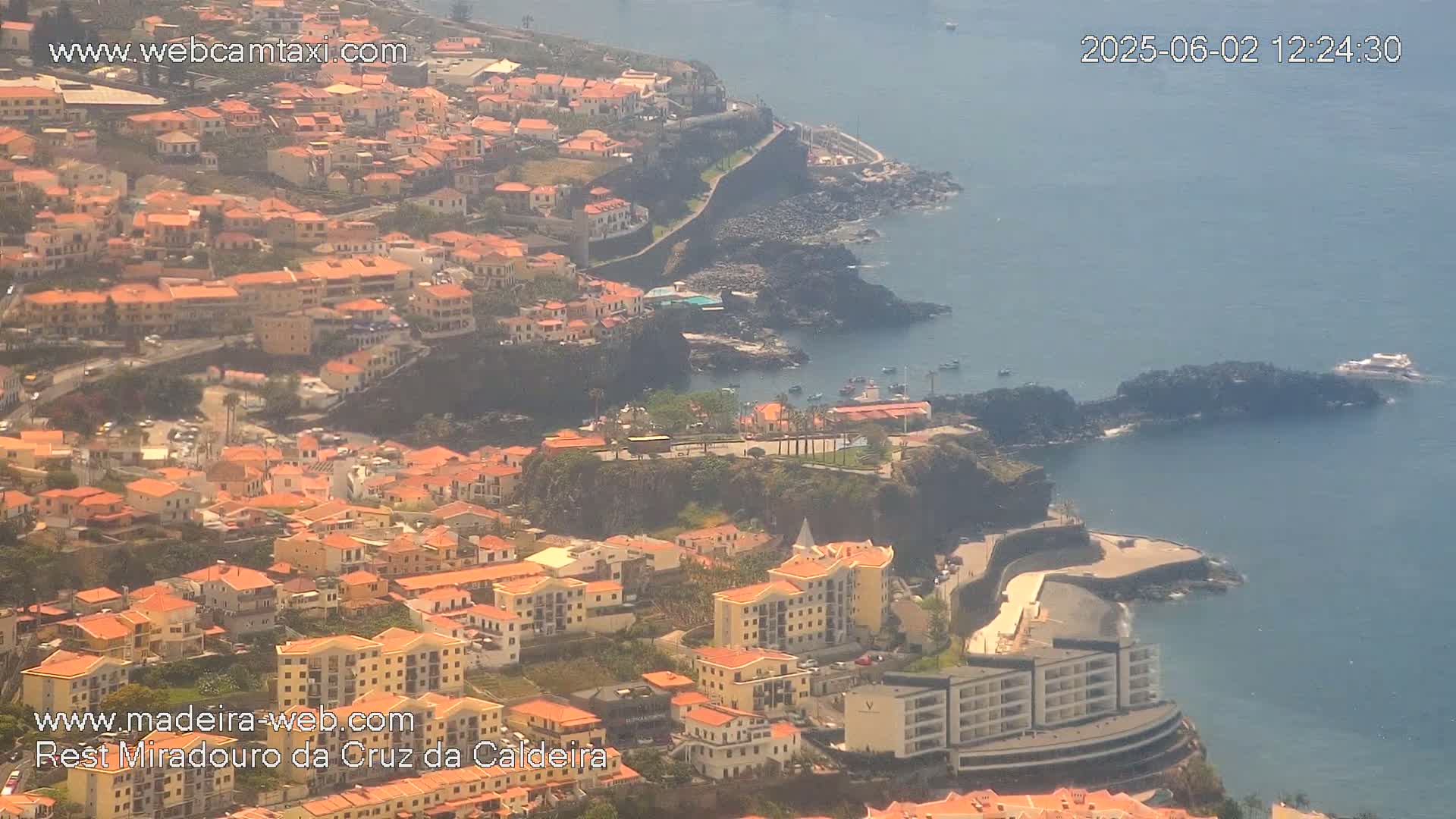 An aerial view shows a coastal town with numerous orange-roofed buildings cascading down a cliffside to a calm ocean on a hazy day.