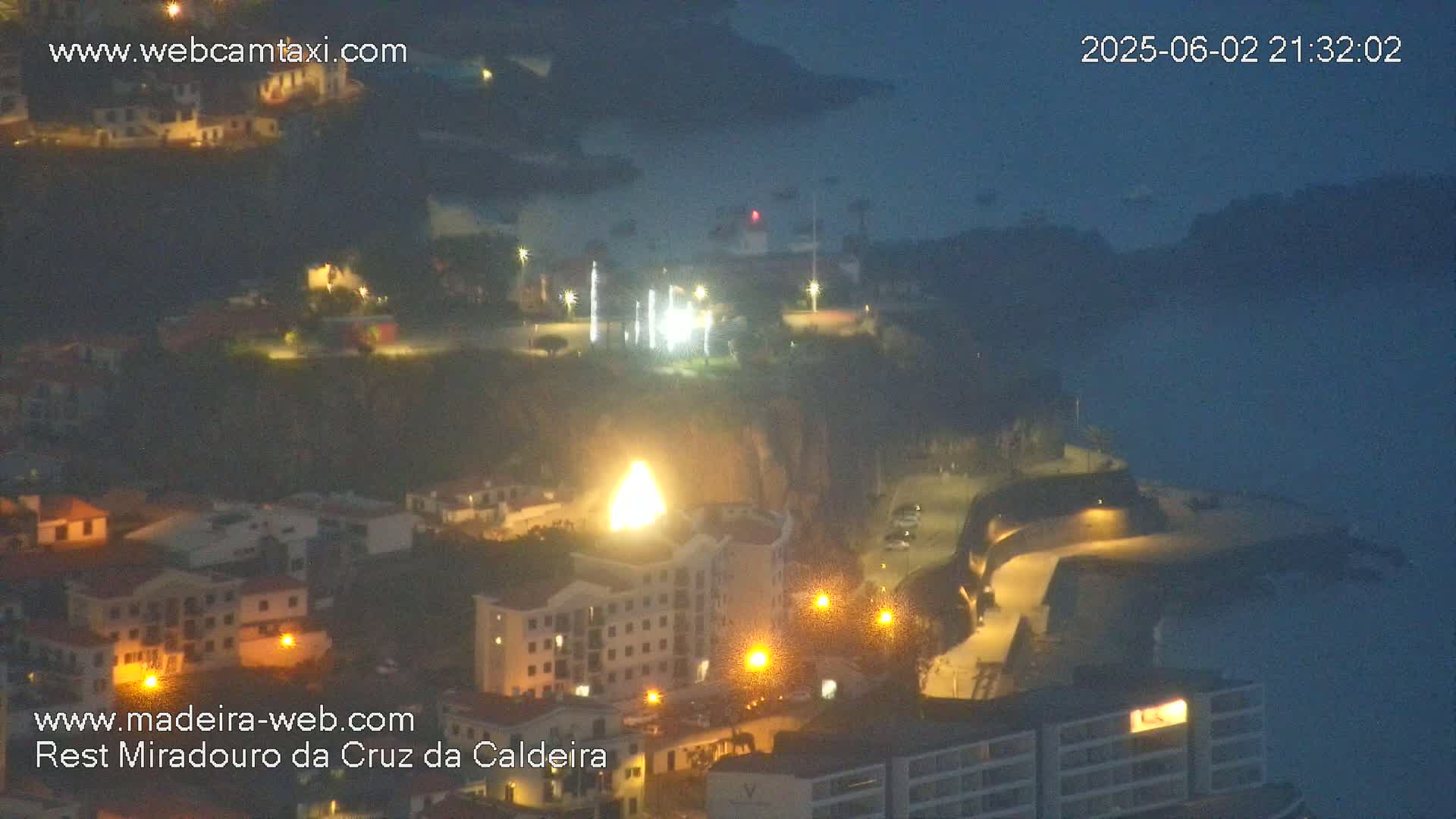 A nighttime coastal town with illuminated buildings and a bright light source near a cliff, overlooking a dark body of water under a mostly clear night sky.
