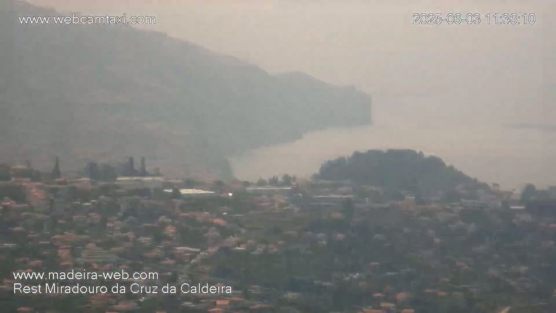 A hazy, long-range view shows a coastal town nestled between mountains and a body of water under a smoky or foggy sky.