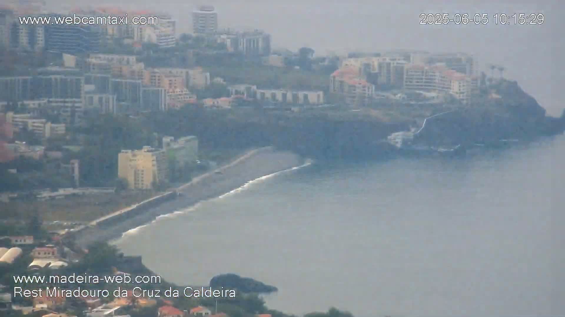 A hazy view shows a coastal town with buildings and a dark-sand beach meeting a calm ocean.
