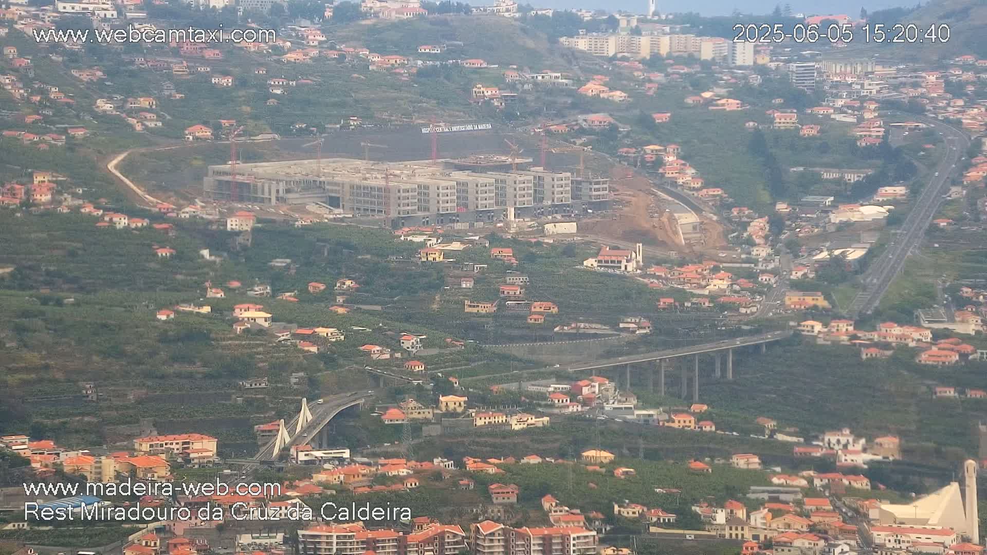 An aerial view shows a hillside town with a large building under construction, a long highway bridge, and hazy weather conditions.