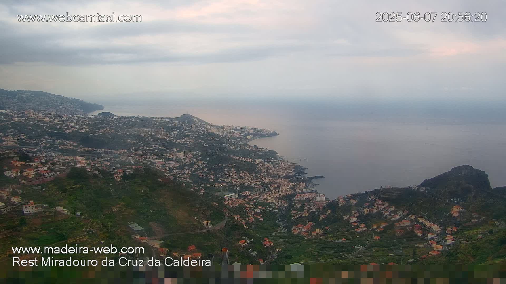 A coastal town sprawls across a hillside overlooking a calm ocean under a cloudy sky.
