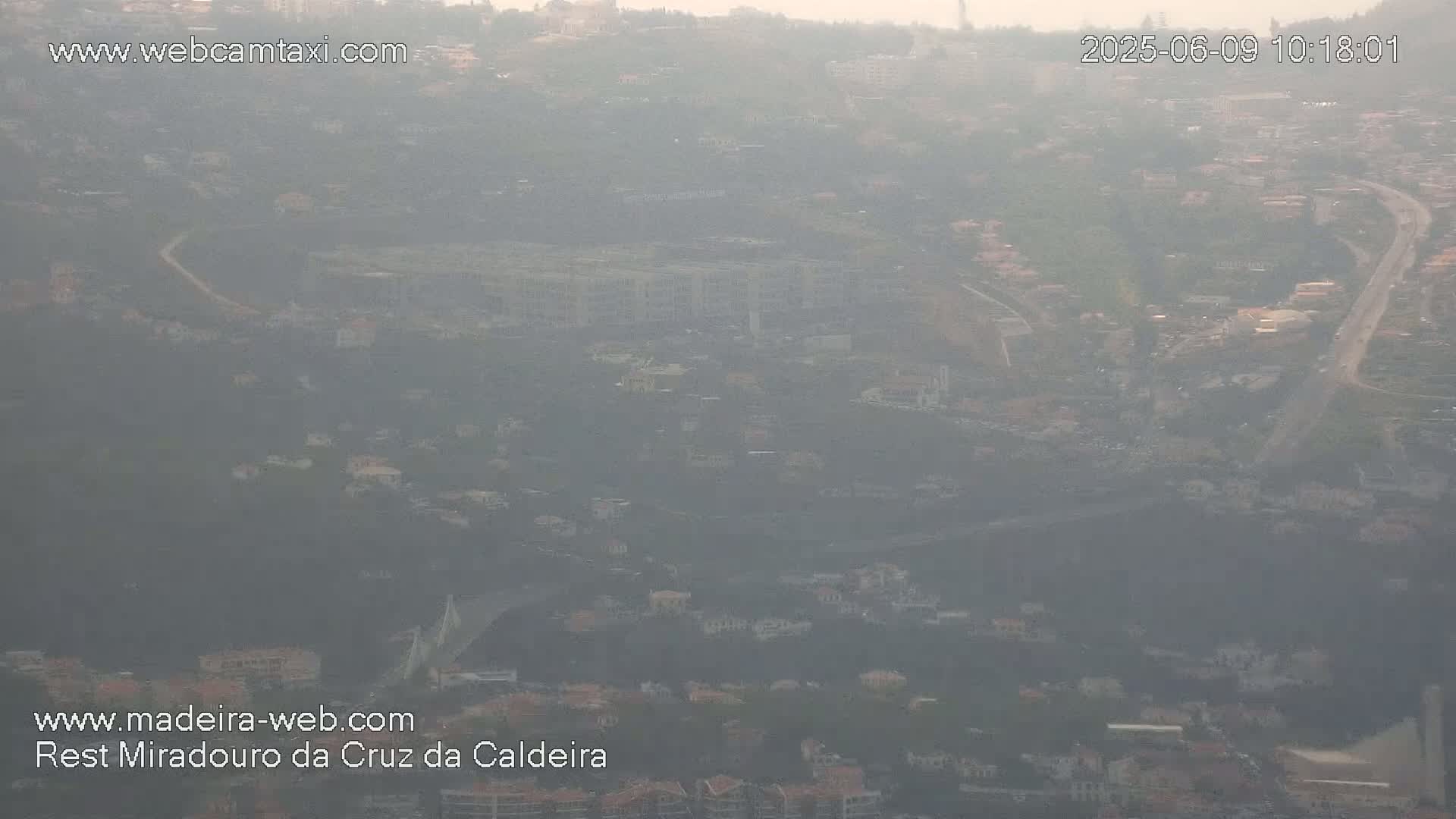 A hazy, high-angle view shows a sprawling residential area with a bridge and buildings partially obscured by atmospheric conditions.