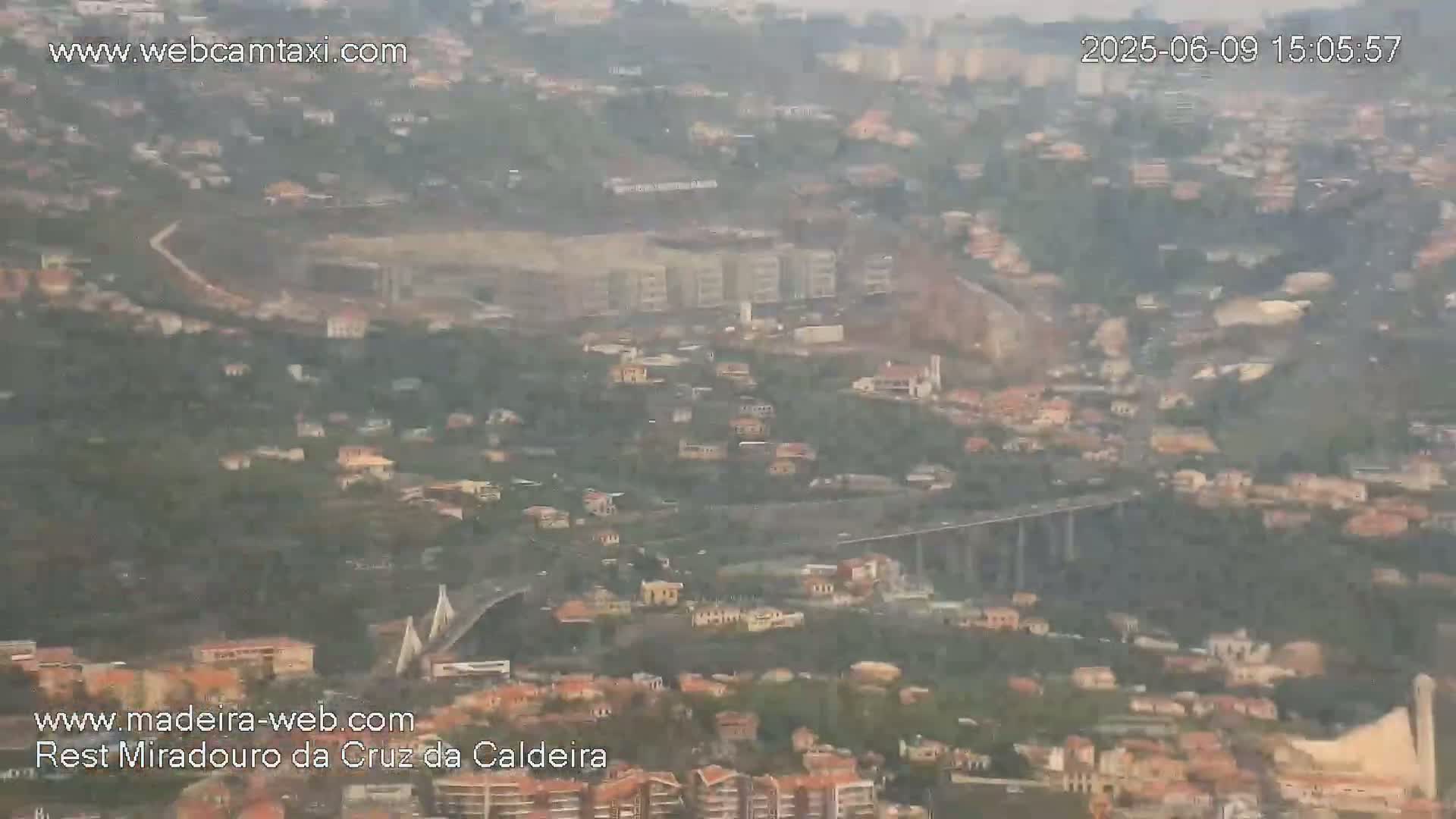 A hazy, long-range view shows a sprawling town nestled in a hillside, featuring a bridge and what appears to be a large stadium or field.