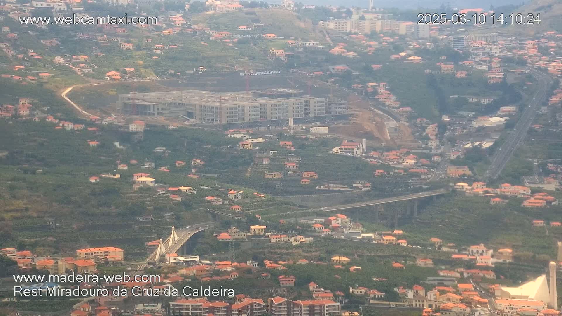 An aerial view shows a hillside town with many low buildings, a large building under construction, and a long elevated highway with a bridge curving through the landscape on a hazy day.