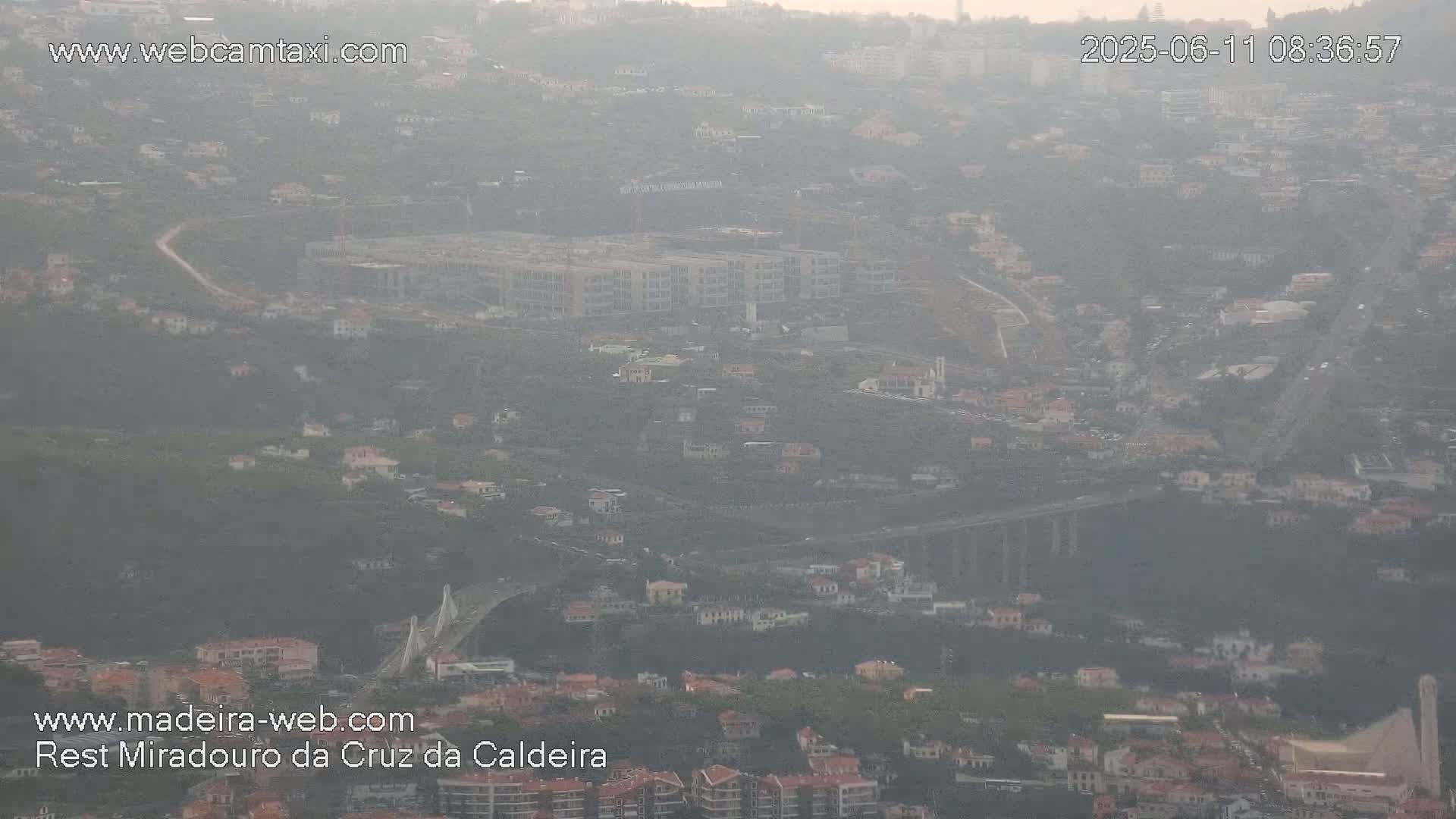 A hazy, high-angle view shows a hillside town with a large building under construction and a bridge visible in the middle ground.