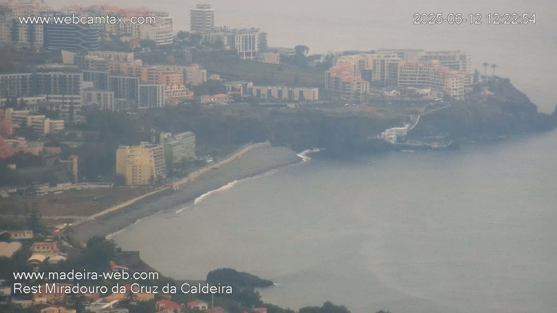 A hazy coastal scene shows a dark-sand beach bordered by buildings and cliffs, with a calm, gray ocean extending to the horizon.