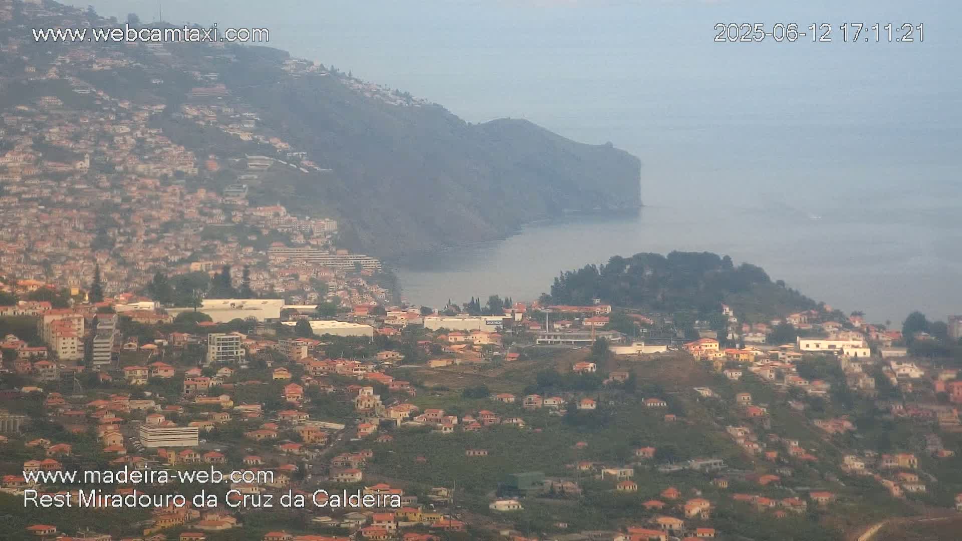 A hazy coastal town sprawls across a hillside, overlooking a calm ocean with a steep cliff in the background.