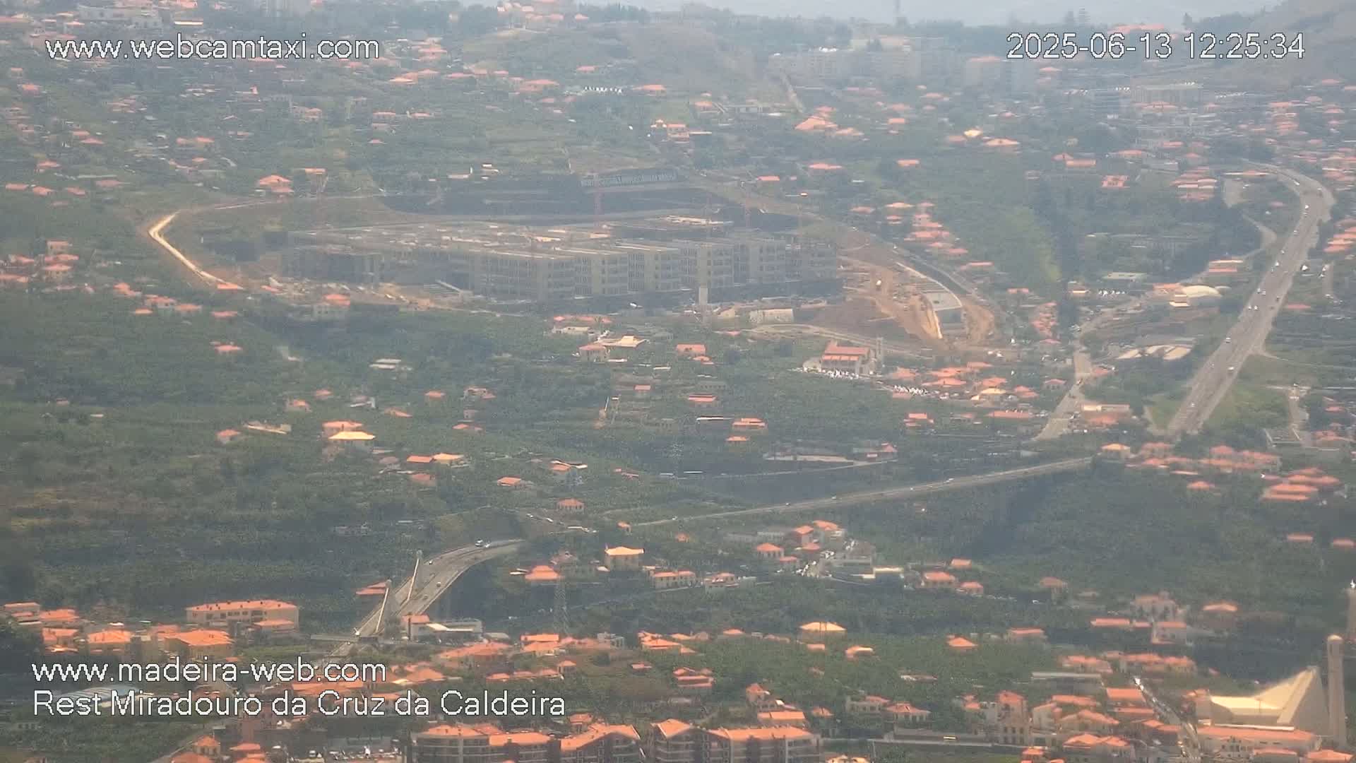 An aerial view shows a large building under construction in a hilly area with many small houses, roads, and lush green vegetation on a hazy day.