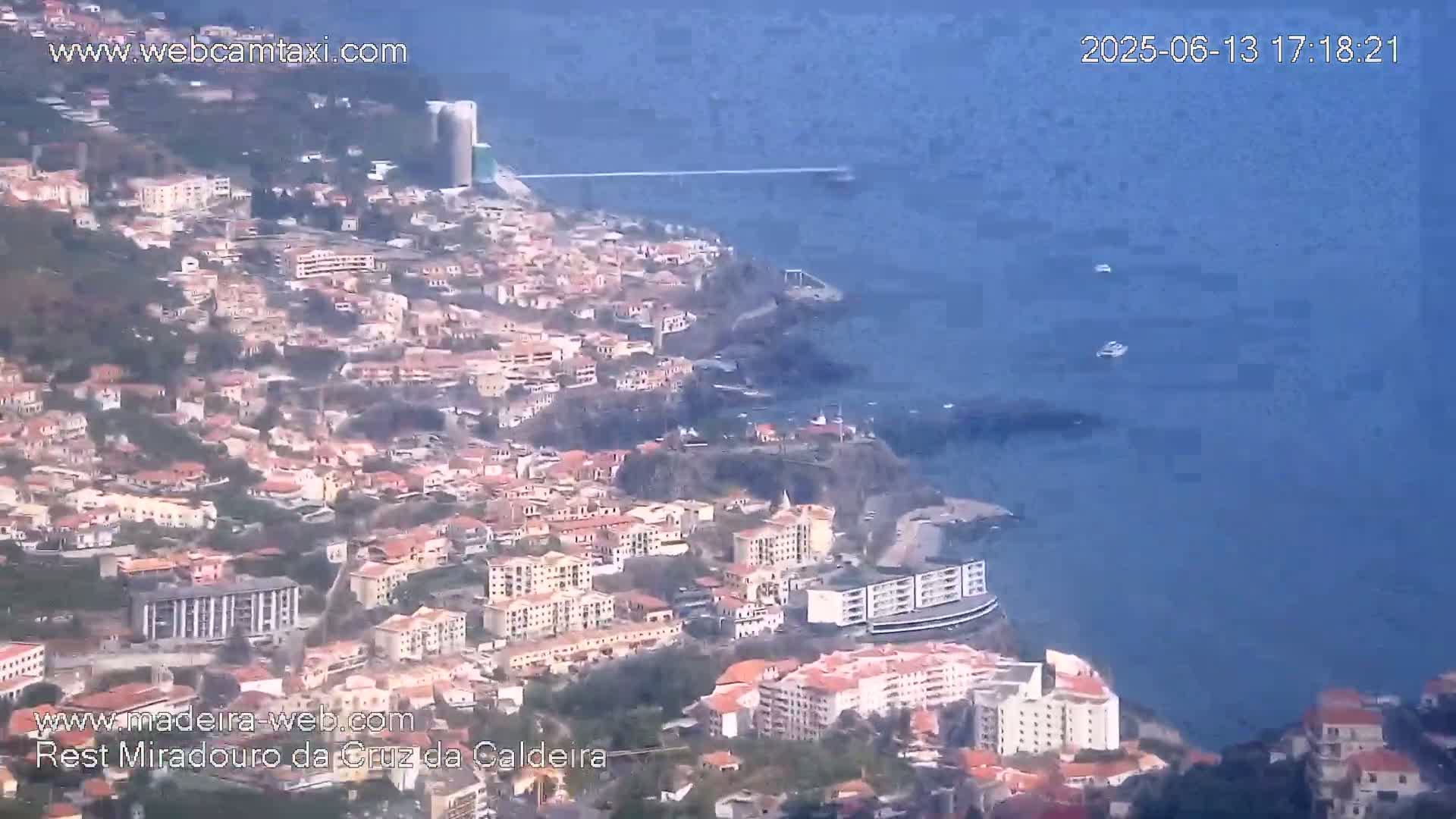 A coastal town with numerous buildings is situated on a hillside overlooking a calm, blue ocean.